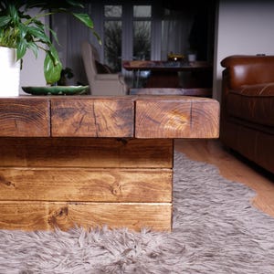 Puede incluir: Una mesa de centro de perfil bajo construida con vigas de madera gruesas te&ntilde;idas de oscuro con patrones de grano y madera de testa visibles. La mesa cuenta con una base robusta en forma de bloque hecha de tablones horizontales.