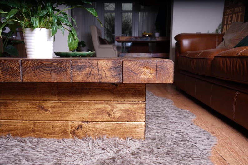 Puede incluir: Una mesa de centro de perfil bajo construida con vigas de madera gruesas te&ntilde;idas de oscuro con patrones de grano y madera de testa visibles. La mesa cuenta con una base robusta en forma de bloque hecha de tablones horizontales.