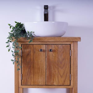 May include: A wooden bathroom vanity cabinet with a natural grain finish, featuring two vertical-plank doors with small round knobs. A white ceramic vessel sink sits on top, paired with a tall chrome faucet and a small green trailing plant.
