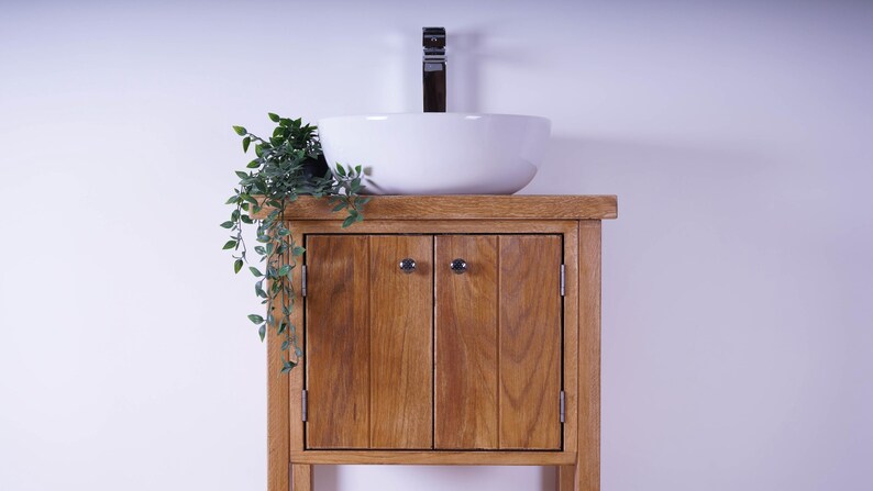 May include: A wooden bathroom vanity cabinet with a natural grain finish, featuring two vertical-plank doors with small round knobs. A white ceramic vessel sink sits on top, paired with a tall chrome faucet and a small green trailing plant.
