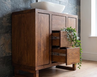 May include: A dark brown wooden bathroom vanity cabinet with a rectangular white ceramic vessel sink and a silver faucet. The vanity features one closed door and two open drawers with metal glides, with a green trailing plant inside the drawers.