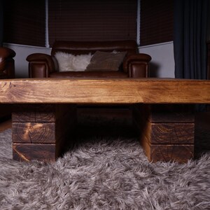 Puede incluir: Una mesa de centro rectangular construida con tablones de madera gruesos te&ntilde;idos de marr&oacute;n oscuro. El tablero presenta una veta horizontal lisa, mientras que la base consta de dos soportes robustos de bloques apilados a cada lado.
