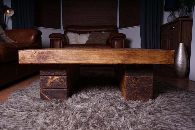 Puede incluir: Una mesa de centro rectangular construida con tablones de madera gruesos te&ntilde;idos de marr&oacute;n oscuro. El tablero presenta una veta horizontal lisa, mientras que la base consta de dos soportes robustos de bloques apilados a cada lado.