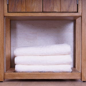 May include: A stack of three folded white towels resting on the lower shelf of a light brown wooden vanity cabinet. The towels are neatly layered, featuring a subtle textured pattern on their surface, positioned within the open wooden frame.