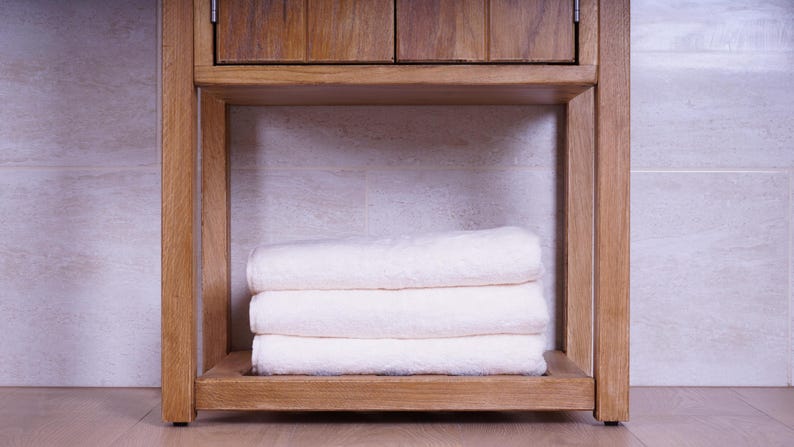 May include: A stack of three folded white towels resting on the lower shelf of a light brown wooden vanity cabinet. The towels are neatly layered, featuring a subtle textured pattern on their surface, positioned within the open wooden frame.