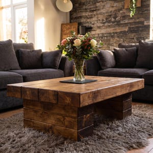 Puede incluir: Una mesa de centro rectangular construida con vigas de madera gruesas te&ntilde;idas de oscuro con una base de bloques de perfil bajo. Un jarr&oacute;n de vidrio transparente con un ramo de rosas blancas y rosas, bayas rojas y hojas de eucalipto encima.