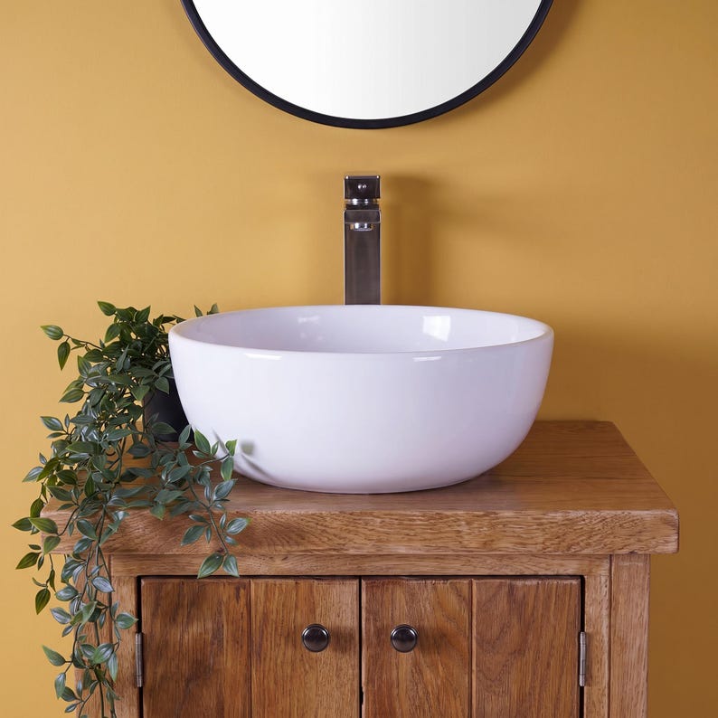 May include: A white ceramic vessel sink with a smooth, rounded basin sits atop a rustic wooden vanity cabinet. A tall, metallic faucet is mounted behind the sink. A green trailing plant is positioned to the left of the basin.