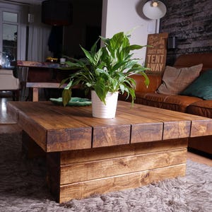 Puede incluir: Una mesa de centro rectangular baja construida con tablones de madera gruesos te&ntilde;idos de marr&oacute;n oscuro con veta visible. La mesa presenta una base maciza en forma de bloque y una superficie plana. Una planta verde est&aacute; en una maceta blanca.