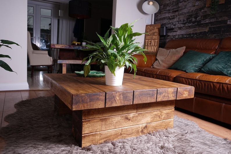 Puede incluir: Una mesa de centro rectangular baja construida con tablones de madera gruesos te&ntilde;idos de marr&oacute;n oscuro con veta visible. La mesa presenta una base maciza en forma de bloque y una superficie plana. Una planta verde est&aacute; en una maceta blanca.