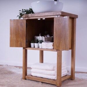 May include: A wooden bathroom vanity cabinet with a white ceramic vessel sink on top. The cabinet features two open doors revealing interior storage with small potted plants and folded white towels, plus a lower shelf holding three stacked white towels.