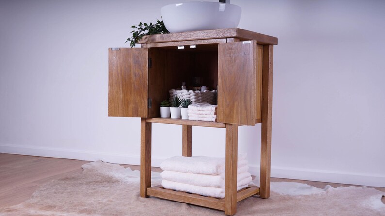 May include: A wooden bathroom vanity cabinet with a white ceramic vessel sink on top. The cabinet features two open doors revealing interior storage with small potted plants and folded white towels, plus a lower shelf holding three stacked white towels.