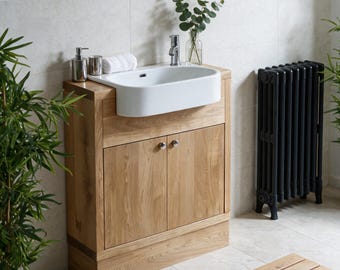 Puede incluir: Un mueble de baño de roble claro con dos puertas con pomos plateados y un lavabo de cerámica blanca semiencastrado. El mueble incluye un grifo cromado, un dispensador de jabón plateado, un vaso y una toalla blanca enrollada en la parte superior.