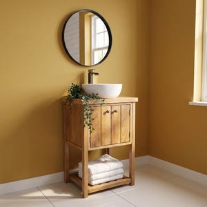 May include: A wooden bathroom vanity with a white vessel sink, a dark metal faucet, and two cabinet doors. A round black-framed mirror hangs above the vanity. A stack of white folded towels rests on the bottom shelf, and a green plant sits on top.