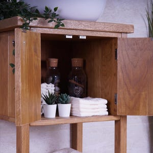 May include: A light brown wooden bathroom vanity cabinet with two open doors revealing an interior shelf. Inside are two glass bottles with cork stoppers, two small potted artificial green plants in white pots, and a stack of folded white towels.