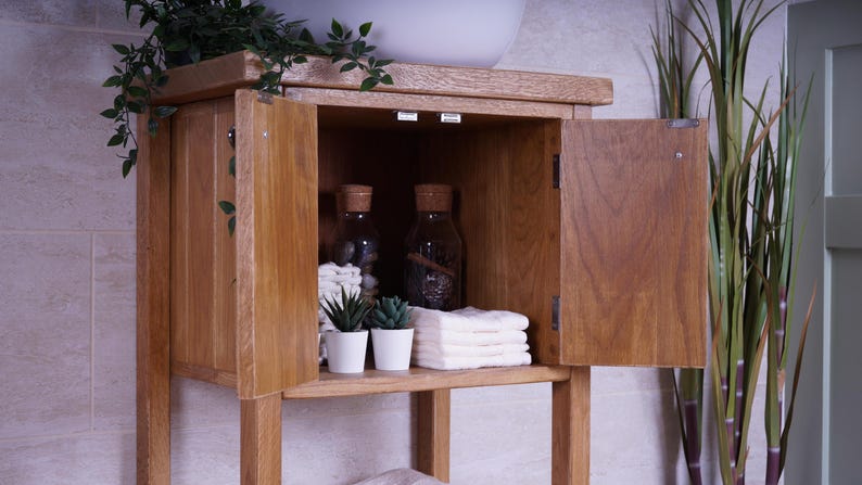May include: A light brown wooden bathroom vanity cabinet with two open doors revealing an interior shelf. Inside are two glass bottles with cork stoppers, two small potted artificial green plants in white pots, and a stack of folded white towels.