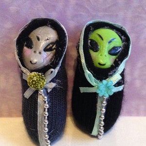 May include: Two small alien dolls wrapped in black fabric with silver beads and blue and green ribbon accents. The aliens have green and gray faces with black eyes.