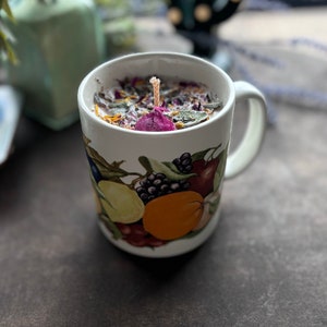 May include: A white ceramic mug with a fruit design, filled with a soy wax candle and dried flowers. The candle has a single wick and is surrounded by dried rose petals, herbs, and other botanicals.