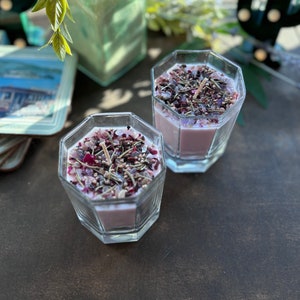 May include: Two pink soy candles in glass jars with a hexagonal shape. The candles are topped with dried flowers, herbs, and crystals.