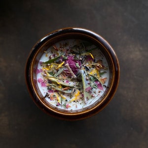 May include: A brown ceramic candle jar with a white wax base and a variety of dried flowers and herbs embedded in the wax. The flowers are pink, yellow, and green.