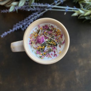 May include: A white candle with dried flowers and herbs embedded in the wax, in a beige ceramic mug with a handle.