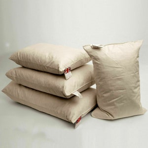 May include: Four white pillows stacked on top of each other. The pillows are made of a soft, natural material and have a slightly textured surface. The top pillow has a small tag with a Union Jack flag on it.