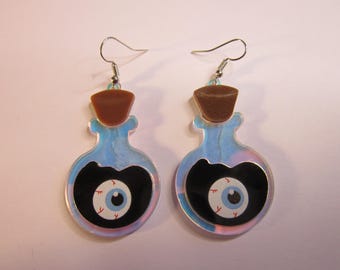 Peut inclure: Une paire de boucles d'oreilles fantaisie en forme de flacons de potion. Chaque boucle d'oreille représente une bouteille bleue avec un bouchon marron et un ovale noir contenant un globe oculaire de dessin animé avec un iris bleu et des veines rouges.