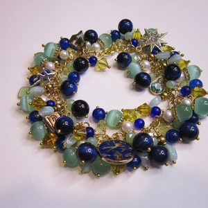 Könnte beinhalten: Ein mehrreihiges Armband mit blauen, grünen und goldenen Perlen. Das Armband verfügt über eine Vielzahl von Anhängern, darunter einen Stern, ein kleines Buch und einen runden Anhänger mit einem Design.