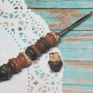 May include: A crochet hook with a silver handle and a brown, white, and black cupcake charm. The hook is lying on a white doily with a brown and blue background.