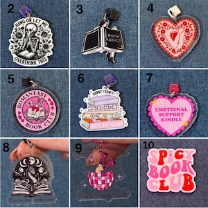 May include: A collection of acrylic charms with various book-themed designs. The charms feature phrases like "Book Addict," "Romantasy Book Club," and "Emotional Support Kindle." Colors include pink, black, white, and red. Some charms have text, and others feature illustrations.