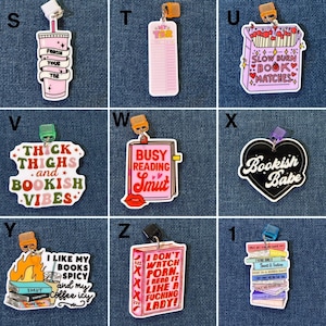 May include: A collection of colorful, whimsical book charms with various designs and text. The charms include a pink drink with text, a pink TBR list, a matchbox with text, a heart-shaped charm with text, a charm with text and a book, a charm with text and a stack of books, and a charm with text and a book with a lip.