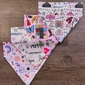 May include: Four triangular dog bandanas with various patterns and text. The designs include references to music, featuring images of guitars, butterflies, and the text "Midnights," "Lover," "Fearless," and "The Eras Tour." The bandanas are on a wooden surface.