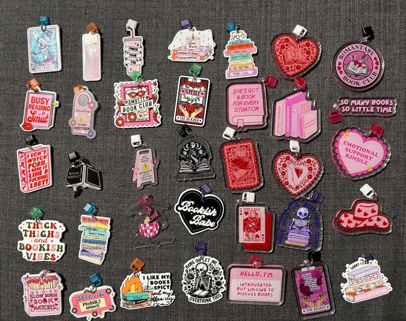 May include: A collection of acrylic book charms with various designs and text. Charms include hearts, books, and phrases like "Bookish Babe" and "Emotional Support Kindle." Colors include pink, red, and black.