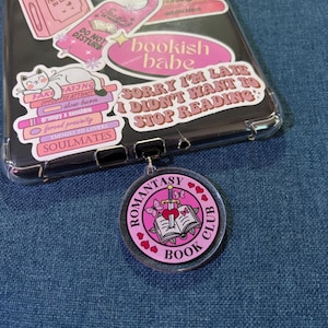 May include: A clear phone case with pink stickers, including a cat, books, and text that reads "bookish babe" and "Sorry I'm late I didn't want to stop reading." A pink "Romantasy Book Club" keychain is attached.