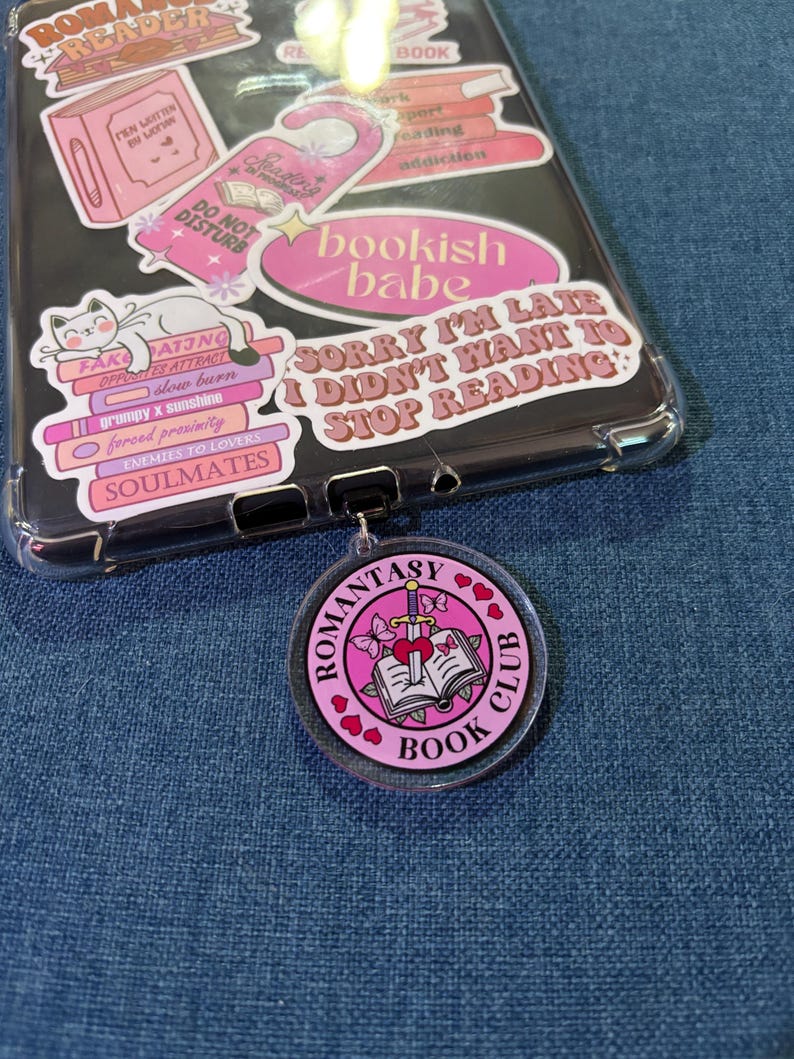 May include: A clear phone case with pink stickers, including a cat, books, and text that reads "bookish babe" and "Sorry I'm late I didn't want to stop reading." A pink "Romantasy Book Club" keychain is attached.