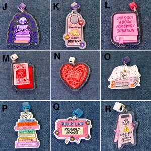 May include: Collection of acrylic charms with various designs. Includes a skeleton reading, a door hanger with "Do Not Disturb", a playing card, a heart with books, a stack of books with a candle, a speech bubble with "Probably Anxious", and a caution sign.