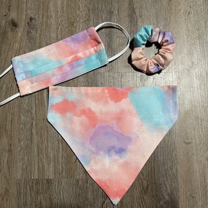 May include: A set of three tie-dye fabric accessories in pink, blue, and purple. The set includes a face mask, a hair scrunchie, and a bandana.