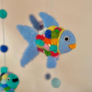 Könnte beinhalten: Handgefertigtes Mobile aus Filz mit einem blauen Fisch. Der Fisch hat einen blauen Körper, Flossen und einen Schwanz. Der Körper ist mit bunten Quadraten und Pailletten verziert. Das Mobile enthält blaue und grüne Filzkugeln.