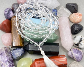 Puede incluir: Un collar mala de cristal blanco con un borla blanca, rodeado de varios cristales y piedras de colores.