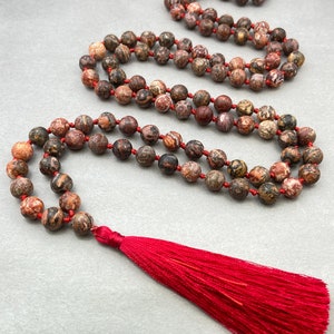 Puede incluir: Un collar de cuentas con una borla roja. El collar presenta cuentas redondas y moteadas en tonos marrones, rojos y beige, ensartadas en un cordón rojo. La borla es de un rojo vibrante, agregando un toque de color al diseño.