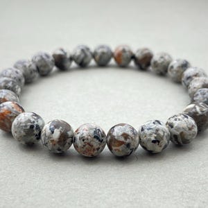 Peut inclure: Un bracelet de perles rondes et mouchetées dans les tons de gris, de marron et d'orange. Le bracelet est fait de pierre naturelle et est monté sur un cordon élastique. Les perles mesurent environ 1 cm de diamètre.