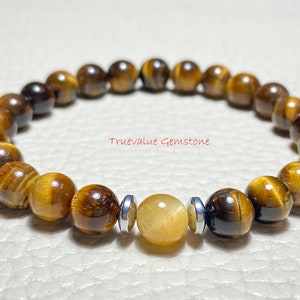 Puede incluir: Un brazalete de cuentas de ojo de tigre marrón y dorado con una sola cuenta de piedra preciosa amarilla y detalles metálicos plateados. El brazalete está sobre una superficie blanca. Truevalue Gemstone.