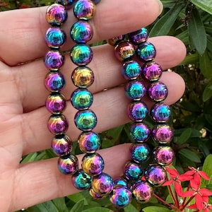 Puede incluir: Dos pulseras de cuentas iridiscentes con un brillo de arcoíris. Las cuentas redondas reflejan colores como el morado, el azul, el verde y el dorado. Las pulseras se sostienen sobre un fondo de follaje verde y flores rojas.