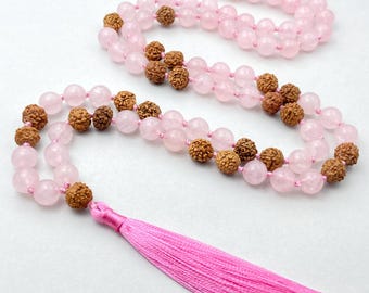 May include: A pink and brown mala necklace with a pink tassel. The necklace is made of rose quartz and rudraksha beads.