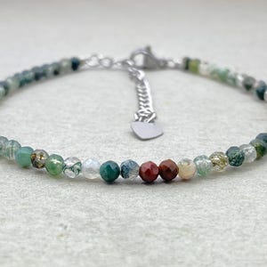 May include: A delicate bracelet made of small, faceted beads in shades of green, brown, and clear. It has a silver-toned chain and clasp, with a small heart charm. A pretty piece of jewellery.
