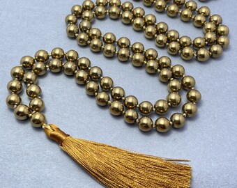 May include: A gold-colored beaded necklace with a tassel. The necklace is made of small, round beads strung together. The tassel is made of gold-colored threads and is attached to the end of the necklace. The necklace is displayed on a light gray surface.