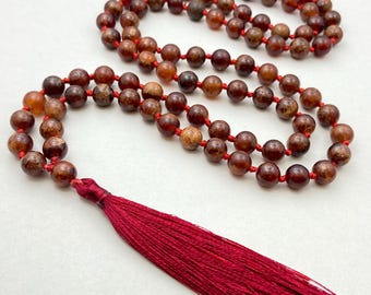 May include: A mala necklace featuring reddish-brown beads strung on a red cord, with a long, matching red tassel. The beads have a natural, speckled appearance. The necklace is displayed on a white surface.