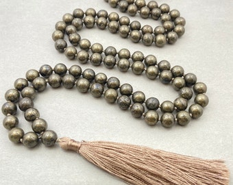 May include: A gold-toned pyrite mala necklace with a tassel. The necklace is made of round beads and has a brown tassel.
