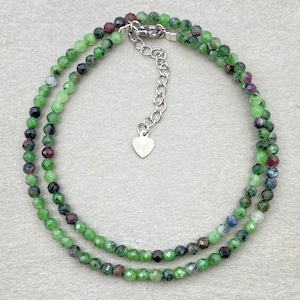 Puede incluir: Un collar de doble cadena hecho de cuentas de piedras preciosas verdes, rosas y azules. El collar tiene un dije de corazón plateado y un cierre plateado.