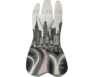 May include: A black and white illustration of a city skyline with the word "ATLANTA" at the top. The city is depicted as a silhouette with a highway in the foreground. The highway is filled with white and red lights.