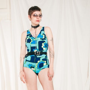 May include: A woman wearing a blue and green patterned swimsuit with a black belt and black knee-high boots. She is wearing sunglasses and a silver chain necklace.